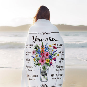 Woman wrapped in a “God says you are” floral blanket standing by the ocean during a calm sunrise — Embrace peace and faith with heartfelt comfort gifts from Hertender at hertender.com.