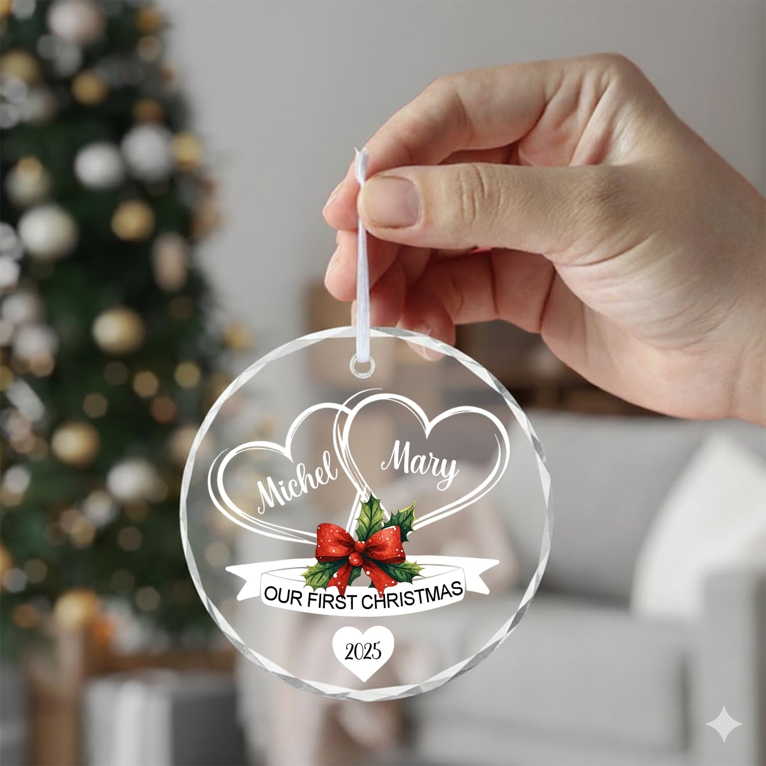 Hand holding a round glass ornament with two hearts and red bow reading “Our First Christmas 2025” near a Christmas tree — Celebrate your love with meaningful personalized Christmas gifts from Hertender.com.