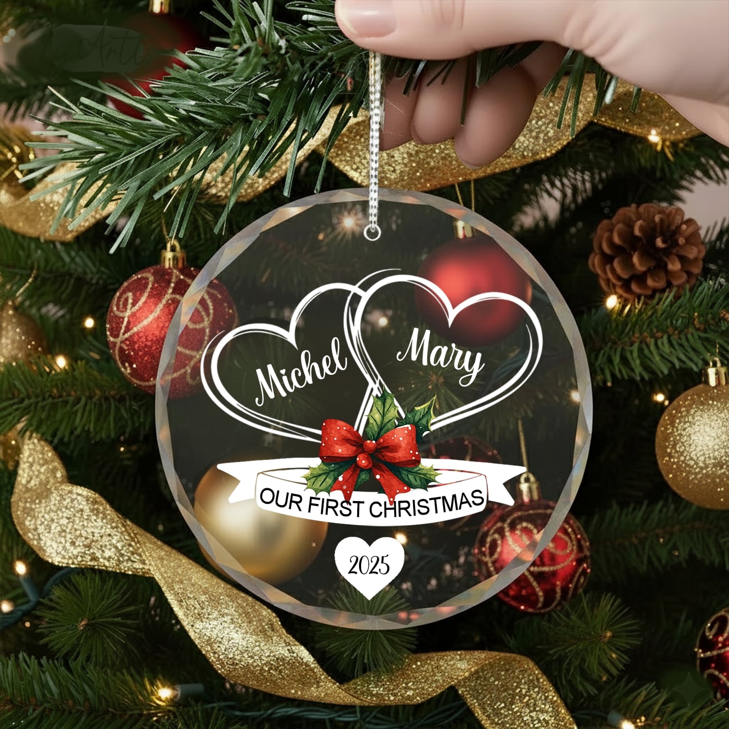 Hand holding a round glass ornament with two hearts and red bow reading “Our First Christmas 2025” on a Christmas tree — Discover heartfelt personalized Christmas gifts at Hertender.com.